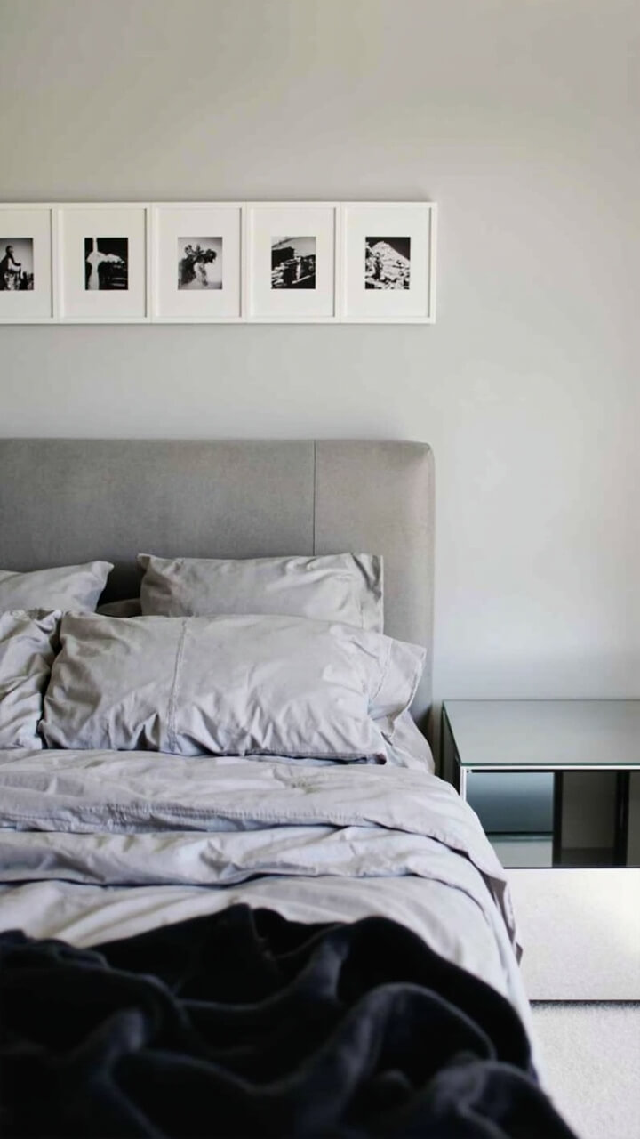Above a soft grey upholstered bed, five matted black-and-white prints in slim white frames form a tight, level arrangement. The nightstand is glossy white, the bedding is monochrome grey, and a black throw sits at the foot.