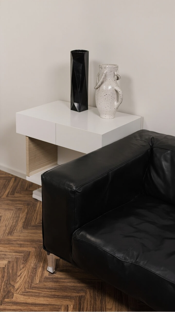 A sleek black leather sofa faces a walnut herringbone rug. Behind it, a minimalist white oak sofa table holds two eye-catching vases: one faceted matte black and one tall speckled white with sculptural handles. No flowers, just the vases themselves.