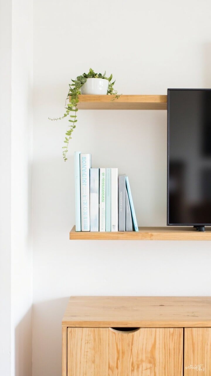 A pale oak wall-mounted shelf above a matching wood TV console, both with gently rounded corners. Bright white walls, a small trailing fern in a white planter, and two rows of color-coordinated books (light blues and greens). No closed storage or visible wires.
