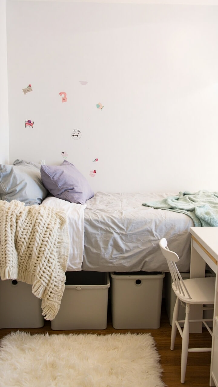 A twin dorm bed nestled against the wall, dressed in a light gray duvet. A chunky ivory knit throw is draped at the foot, with a soft mint fleece blanket folded at the side. A single lavender throw pillow rests between standard dorm pillows. Storage bins peek out under the slightly raised bed. On the wall hang removable pastel graphic decals. There’s a simple rug just under the edge of the bed, and a basic desk and chair nearby.
