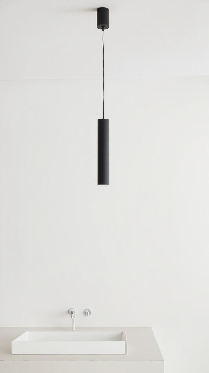 A white ceiling with one single matte black cylindrical pendant hanging directly above a simple rectangular undermount sink. The cord is barely noticeable, and the rest of the vanity is uncluttered.