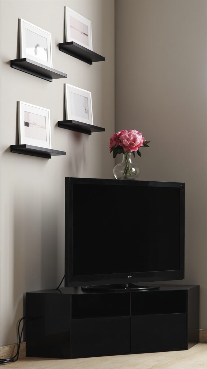 A narrow, black lacquered corner stand supports a 42-inch TV. Above and to the side, three slim picture ledges stagger up the wall, each displaying one or two minimalist art prints in thin white mats. A small clear glass vase with pink peonies rests on the stand. The tv cords are neatly wrangled in a matte black cable channel.