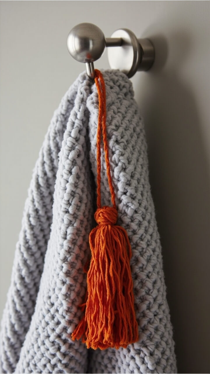 A round brushed nickel hook on the wall holds a chunky gray cotton robe. From the hook, a faux velvet orange pumpkin tassel hangs, adding a dash of color.