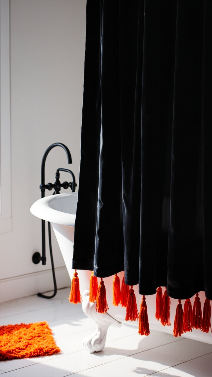 A clawfoot tub is hugged by a thick charcoal black shower curtain, detailed with bright orange tassels along the bottom hem. The curtain hangs from black matte hooks. In the corner, a small orange bath mat sits on white tile floor.