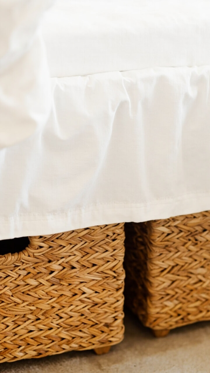 Two low-profile natural rattan baskets are half-visible beneath a platform bed. The baskets’ woven texture shows against a white cotton dust ruffle.