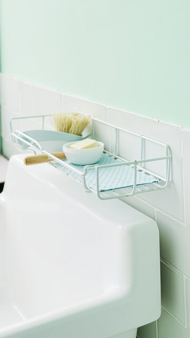 A narrow white-coated wire shelf is mounted above a white porcelain utility sink. On the shelf: a wood-handled scrub brush, small bar soap in a ceramic dish, and a pale blue soaking tray. The wall is pale mint with a thin white tile backsplash.