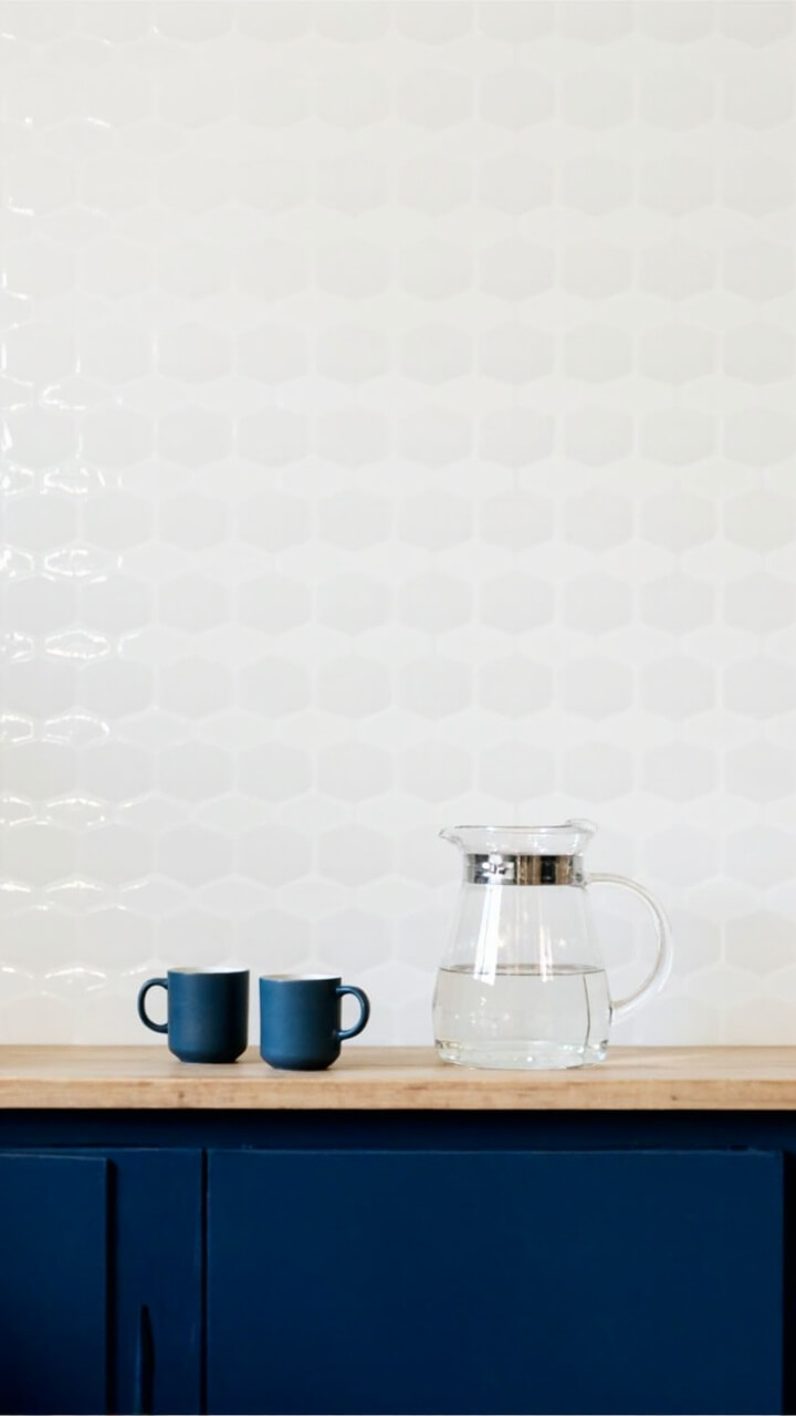 One kitchen wall is covered with matte white elongated hexagon tiles. Integrated beneath it is a floating light-wood shelf holding two matte blue coffee mugs and a glass carafe of filtered water. Lower cabinetry in deep navy adds a bold contrast.