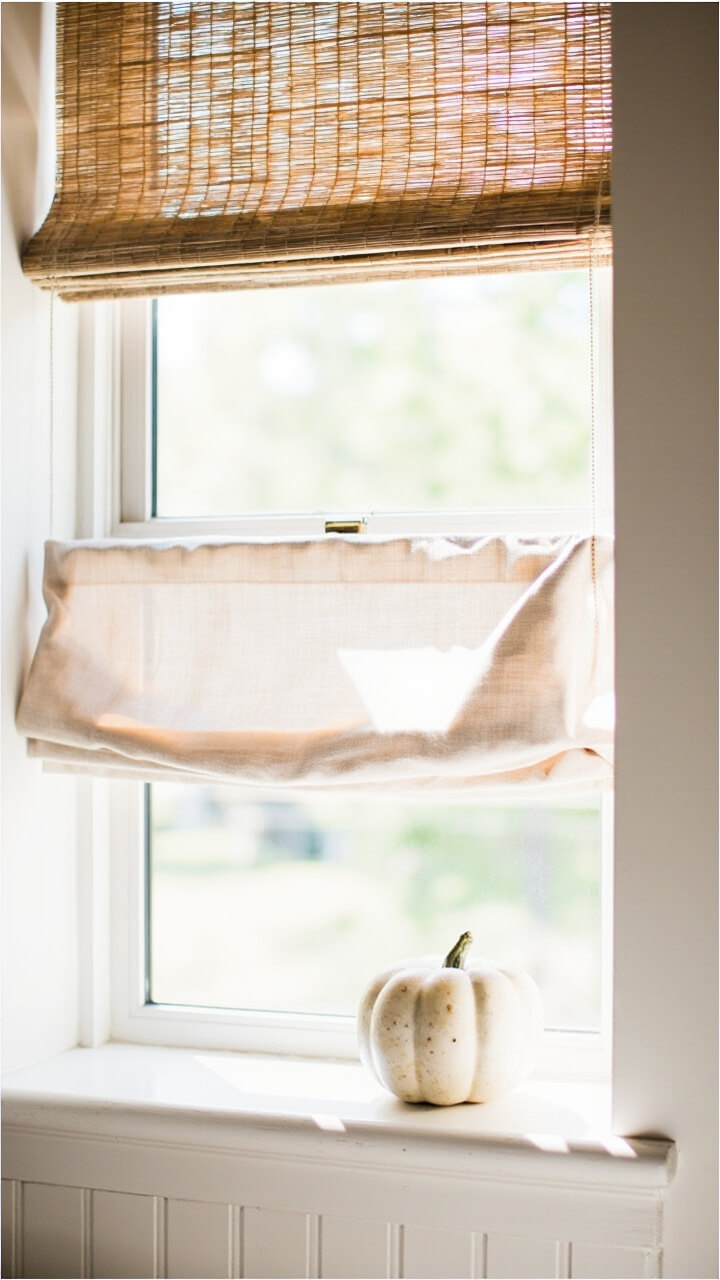 At a small bathroom window a natural bamboo roman shade is mounted inside the frame. An oat linen cafe curtain hangs across the lower half on a matte brass tension rod. A small alabaster ceramic pumpkin rests on the windowsill.
