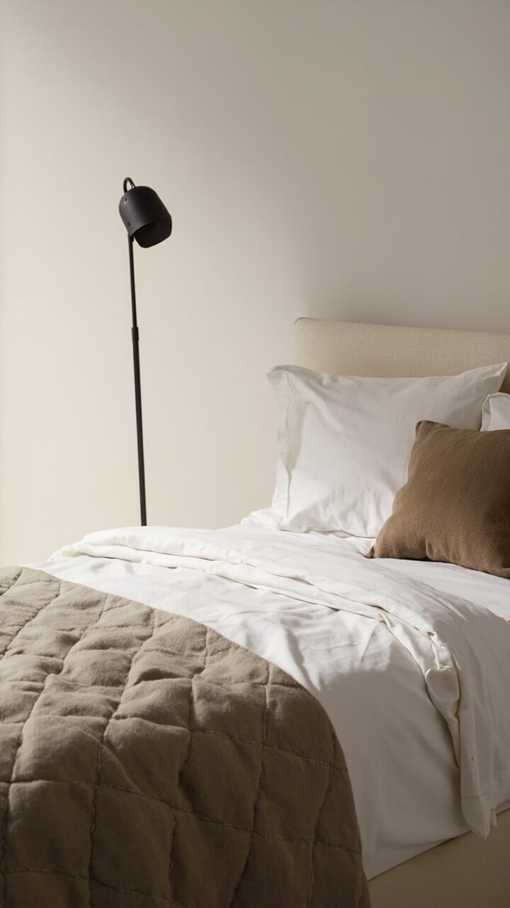 Twin bed with ivory percale sheets, a taupe matelassé coverlet, a beige wool throw at the foot, a single sand-colored accent pillow, and a black metal floor lamp beside the bed.