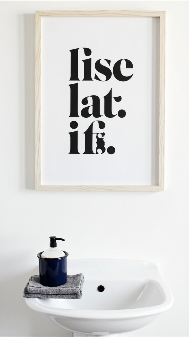 A black and white graphic print in clean sans serif font, framed in pale unfinished wood and hung above a small white utility sink. Beside the sink, a navy blue enamel soap dish and a folded gray washcloth complete the setting.