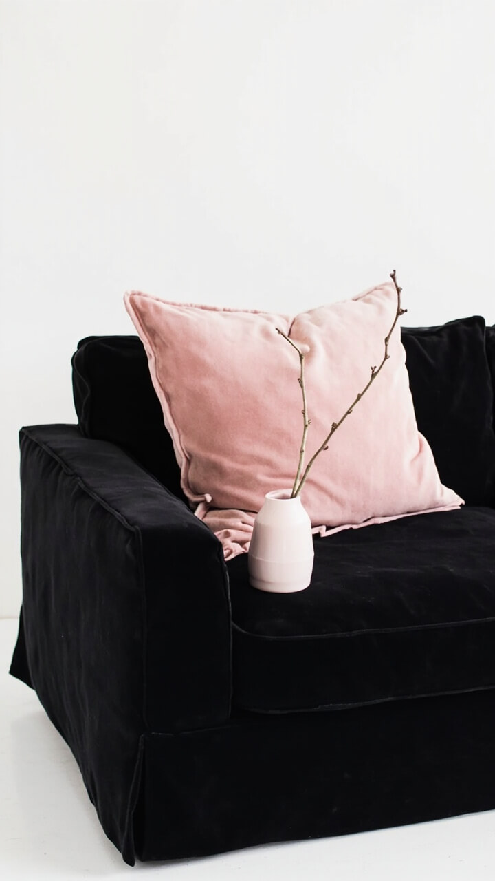 A black sofa anchored by two blush pink velvet pillows. On the arm, a pale blush ceramic vase holds a single branch. The rest is clean and minimal.