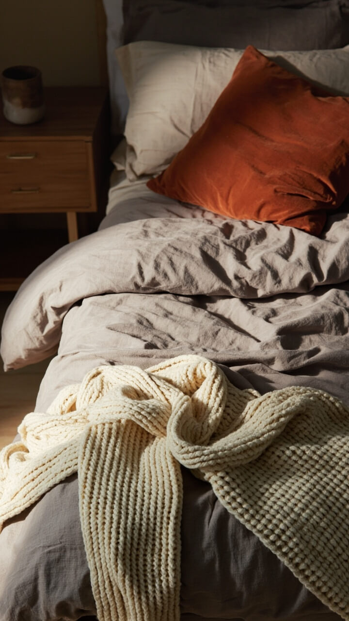 A queen bed with a textured taupe linen duvet, a thick cream chunky knit throw casually draped at the foot, two deep orange velvet pillows, and a simple wooden nightstand beside the bed with a ceramic mug.