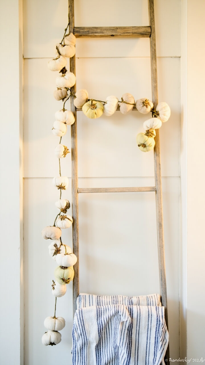 A narrow antique wood ladder leans against white shiplap. A garland of small fabric pumpkins in shades of white, beige, and muted orange is strung on the middle rung. Two folded striped linen hand towels rest on lower rungs.