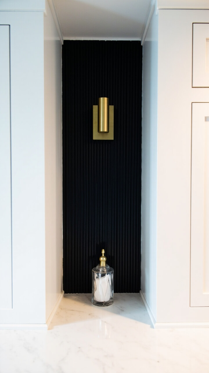 Between two sections of glossy white cabinets, a vertical panel of matte black ridged wall extends from countertop to ceiling. A single brushed brass wall sconce is mounted near the top. The counter below is white marble with a large crystal salt grinder.