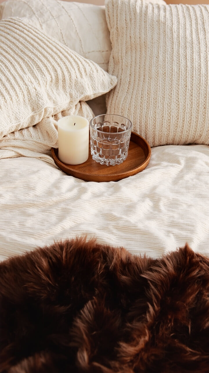 A light oatmeal duvet, a chocolate brown faux fur throw folded at the foot of the bed, a pair of cream knit pillow shams, and a small round wooden tray with a glass candle on the bed.