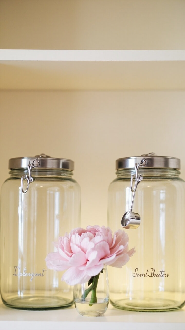 Two large glass apothecary jars with brushed nickel lids labeled “Detergent” and “Scent Boosters” sit clustered on a warm white open shelf. A small silver scoop is hooked on the side of one jar, and a single blush pink peony in a clear bud vase brightens the middle.