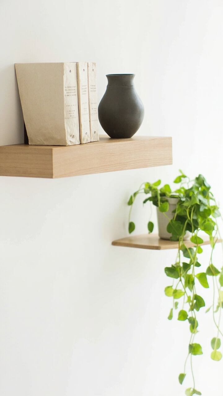 Two light oak floating shelves are stacked on a matte white wall. One shelf holds three cream-colored hardcover books and a charcoal matte vase. The other features a single trailing pothos plant in a white ceramic pot.