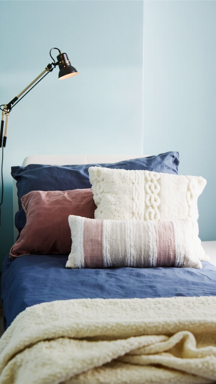 A twin dorm bed against a pale blue wall has three throw pillows: a dusty rose velvet square, an ivory cable-knit rectangle, and a striped blush and white lumbar pillow. The bed has a navy sheet set, and a soft cream blanket is folded at the foot. A reading lamp is clipped to a shelf above.