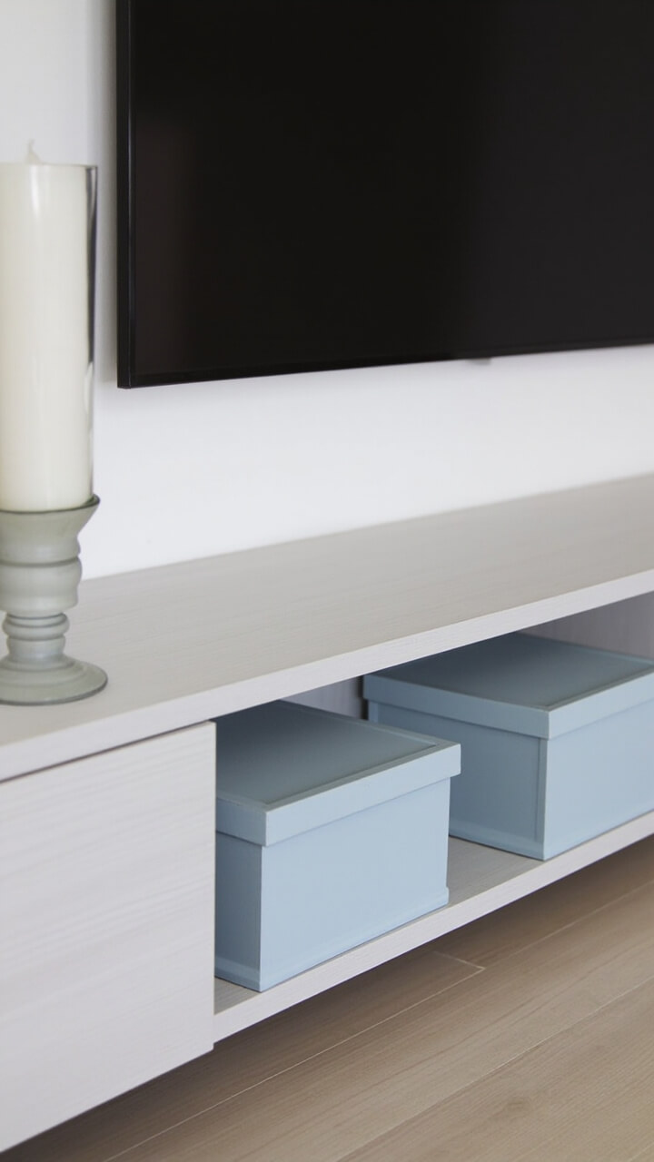 A long, matte pale grey wood TV console, subtly visible grain. Centered beneath the TV, two pale blue storage boxes on the bottom shelf, and a frosted glass hurricane lamp with a white pillar candle sits at the left end.