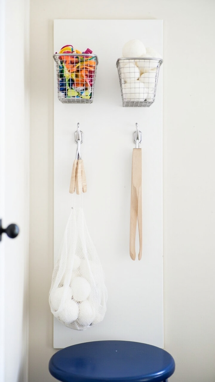 A narrow white pegboard is installed on a side wall, holding two wire baskets (one with sock clips, one with dryer balls), a pair of wooden tongs, and a mesh zip laundry bag. There’s a small navy blue stool below.