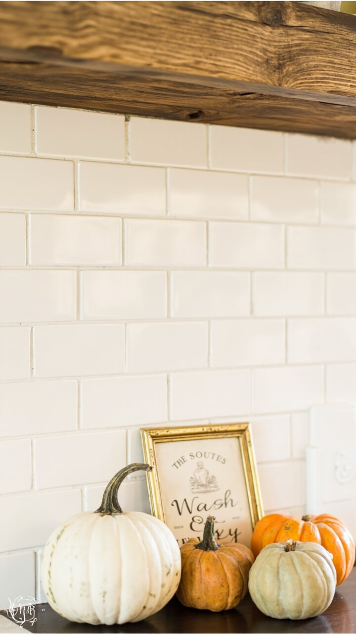 A weathered floating pine shelf above a white subway tile backsplash. On one end: three pumpkins (large off-white, medium tan, small muted orange), leaning against a standing vintage brass framed sign that reads “Wash & Dry.”