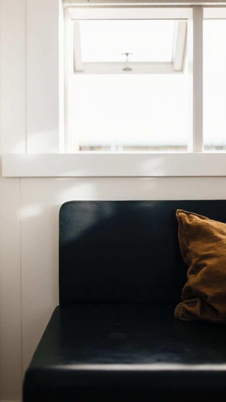 A long, flat black wood bench sits under window light. At one end sits a tan linen lumbar pillow. The rest of the bench is clear, revealing the clean edges and smooth finish.