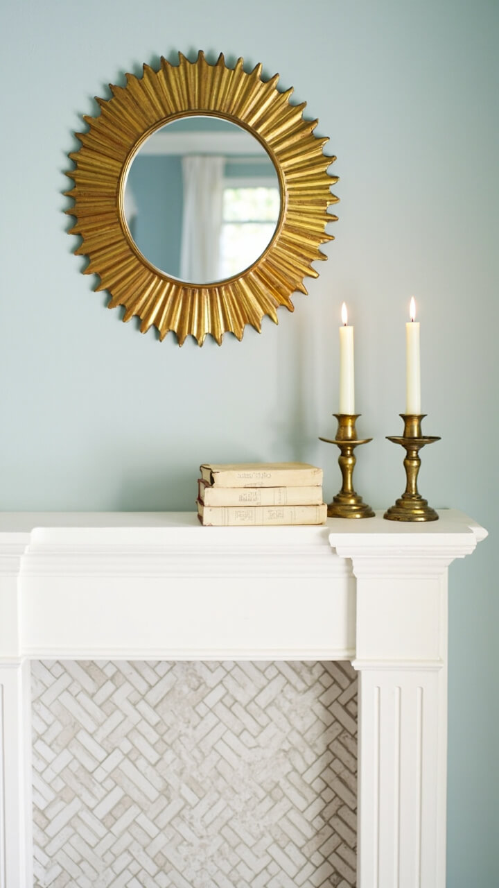 A medium-sized brass sunburst mirror centers above a classic white painted mantel. On the mantel: two matching tapered brass candlesticks with white candles and a stack of cream linen-bound books. There’s a herringbone tile fireplace and pale blue walls in the background.