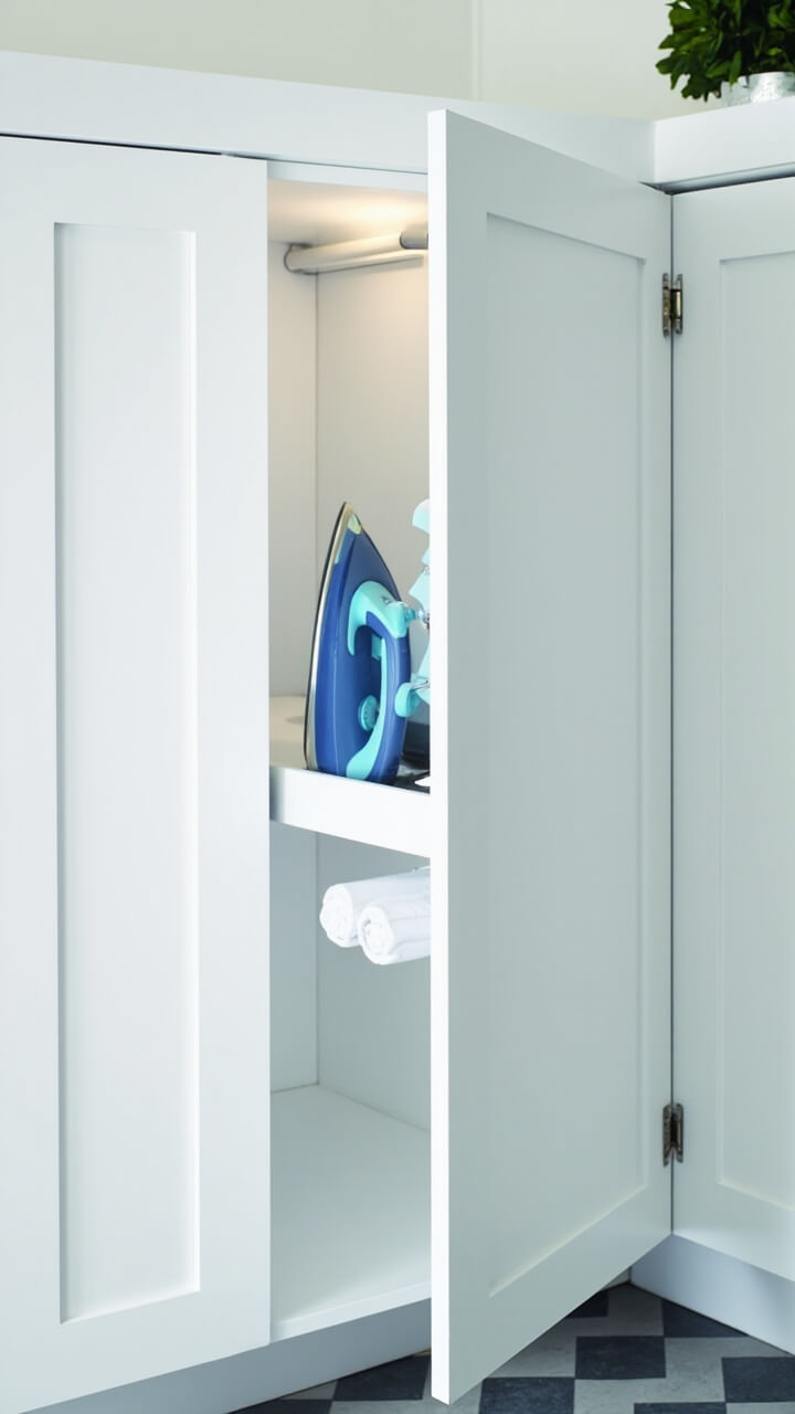 A tall white cabinet with a single pull opens to show a neatly stored folding ironing board, a matte blue iron, and a rolled white ironing cloth. The rest of the nook is bright and uncluttered, with gray and white geometric tile underfoot.