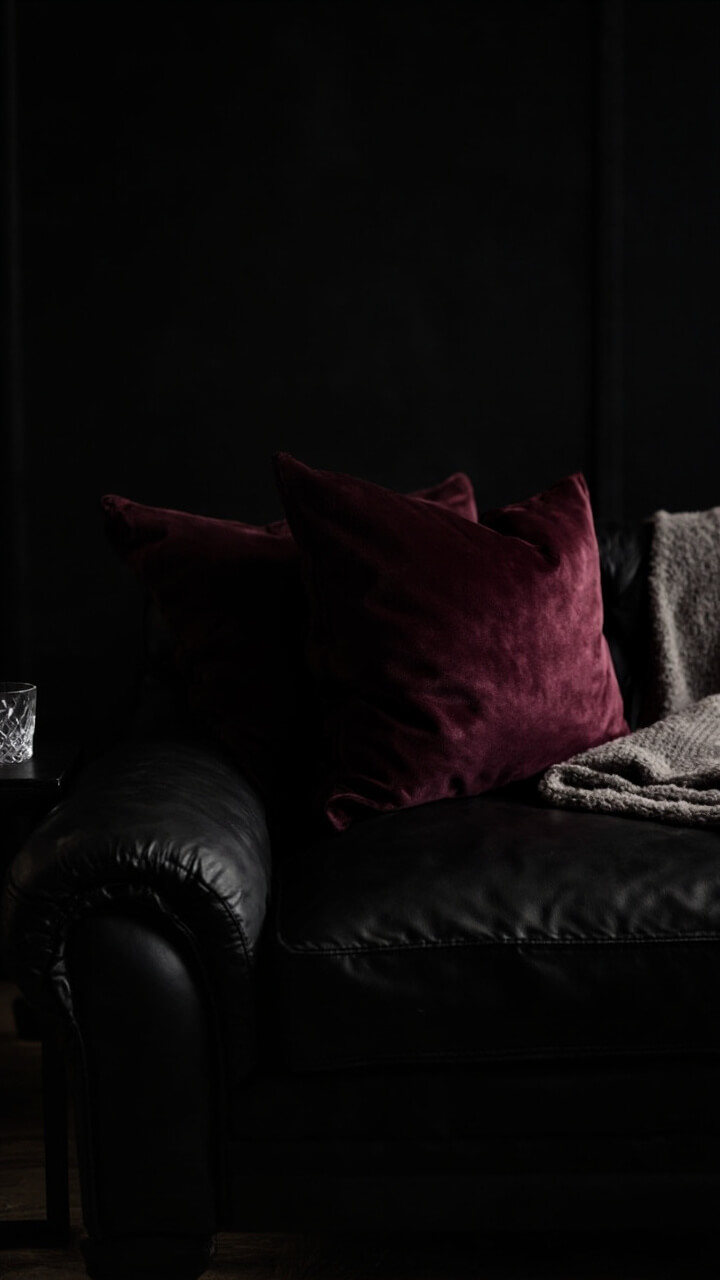 A black sofa holds two burgundy velvet pillows and a folded gray wool blanket. On the side, a small matte black side table has a single crystal whiskey glass.