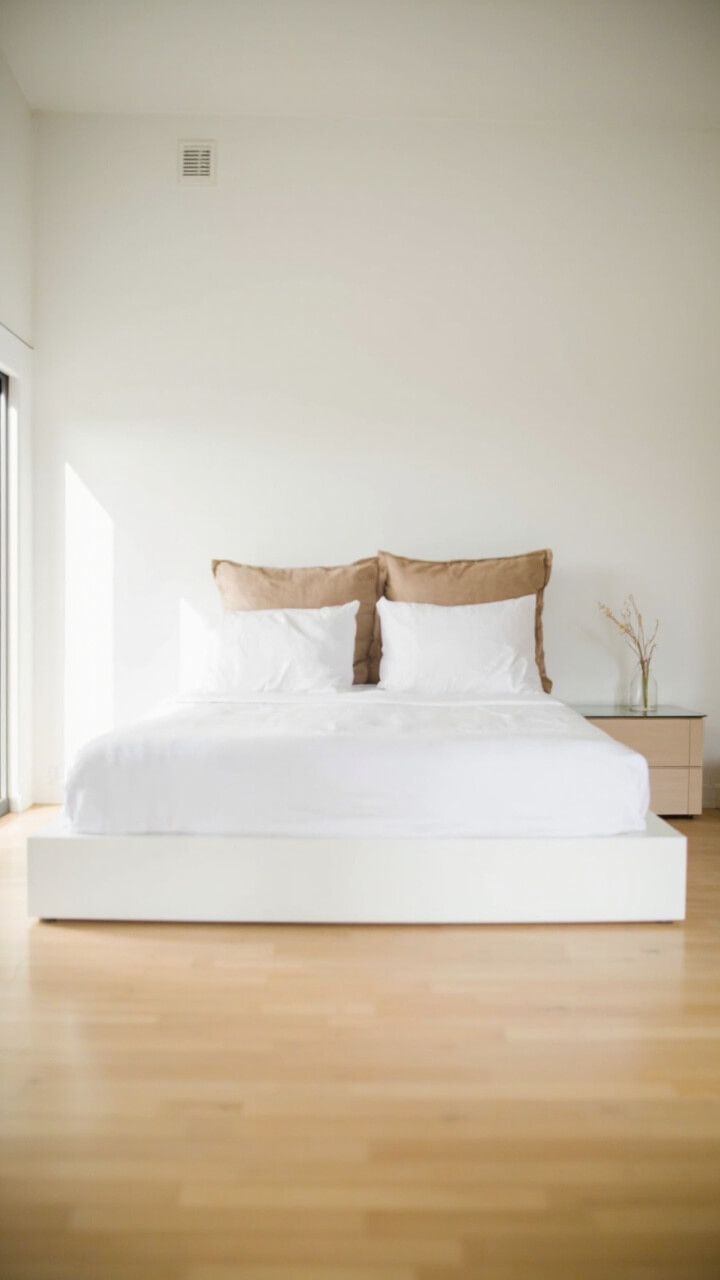 A white platform bed holds an off-white cotton duvet, topped with a light gray knitted wool throw folded across the foot. Two large, sand-colored linen euro shams rest against the headboard. The setup is balanced and airy, surrounded by simple, light oak floors and an uncluttered nightstand.