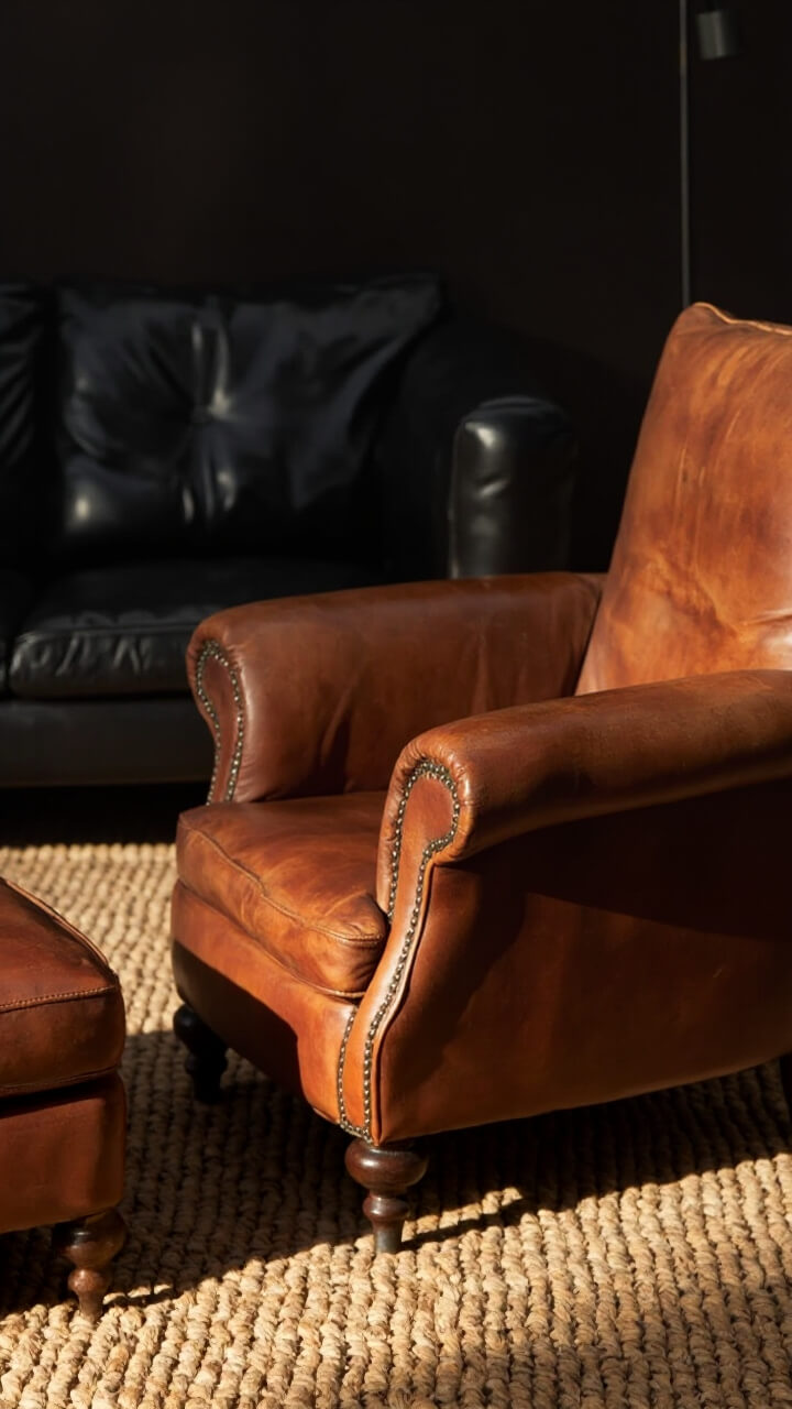 A structured black sofa faces a caramel cognac leather armchair with a matching ottoman. In front, a light tan jute rug adds earthiness.