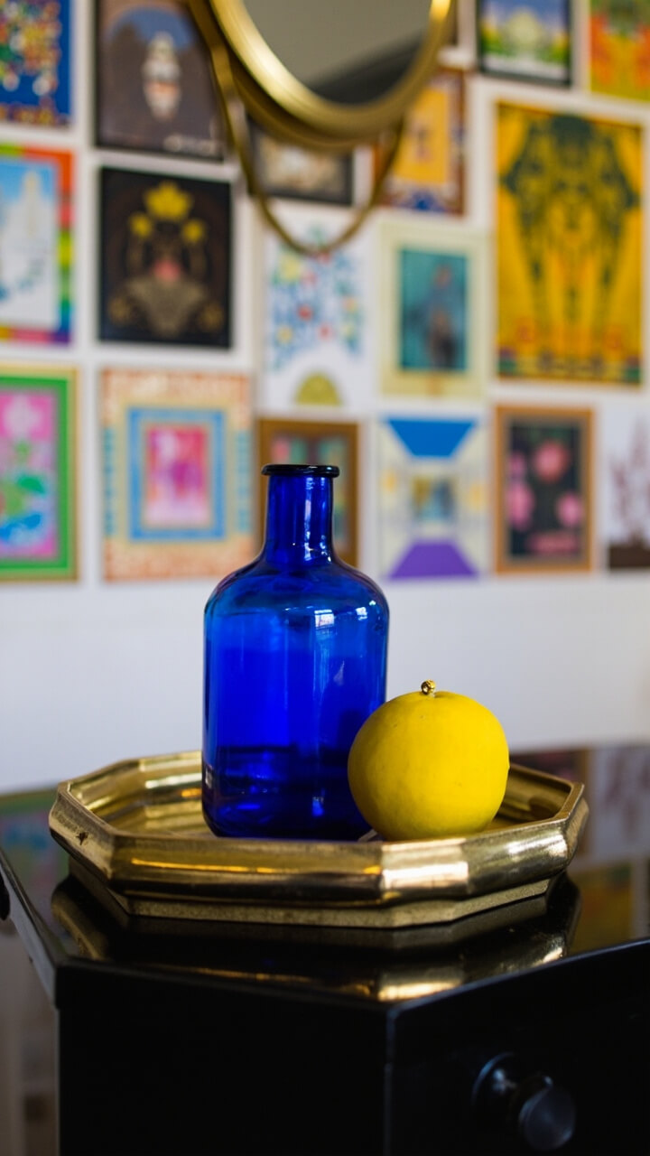 A hexagonal mirror tray with a brass rim sits atop a black lacquer side table. On the tray: a cobalt blue glass bottle vase and a bright yellow ceramic orb. Behind, a wall collage of small, colorful framed art pieces surrounds a round brass wall mirror.