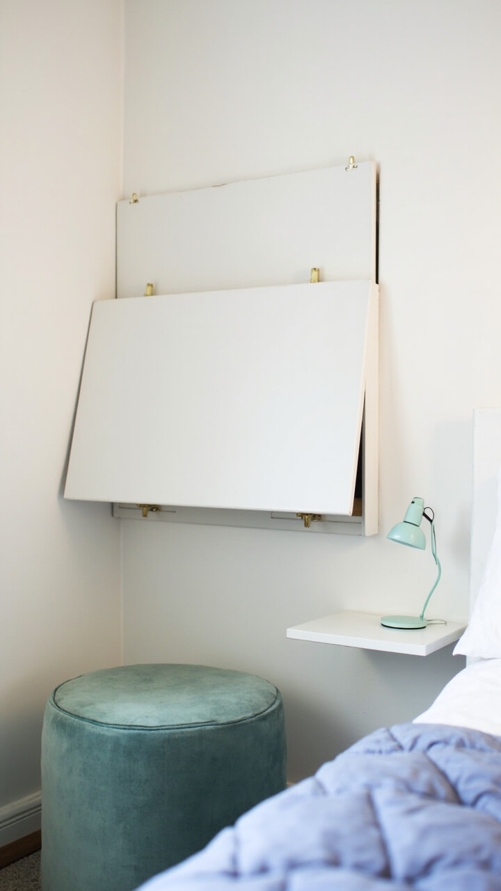 A white wall features a mounted fold-down desk with brass hinges, closed when not in use. Next to the desk, a small mint green reading lamp sits on a petite shelf with a matching mint ottoman tucked under. On the other side of the room is a low twin bed with a periwinkle quilt.