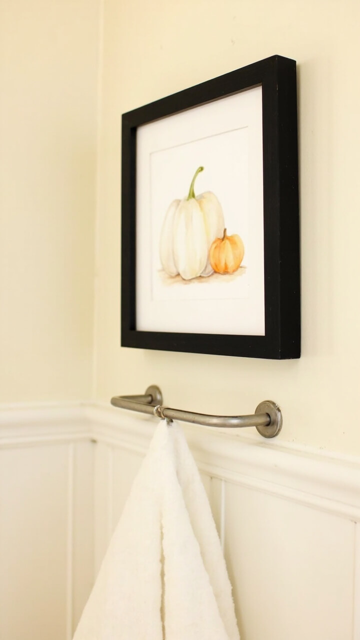 A small black wood frame hangs above the towel bar on a creamy wall. Inside is a minimal illustration of three watercolor pumpkins (white, light orange, and pale green) on a soft background.