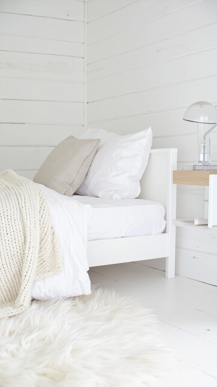 A small white platform bed stands against a white shiplap wall. The bedding is a mix: a chunky ivory knit throw, crisp cotton sheets, and a linen Euro pillow. The nightstand is pale birch wood with a clear glass lamp. The rug is fluffy, soft white.